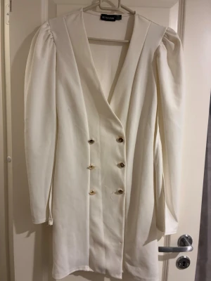 Vit blazerklänning från PrettyLittleThing - Snygg vit blazerklänning från PrettyLittleThing med markerade axlar, djup v-ringning och guldknappar framtill. Klänningen har lång ärm och en rak, kort passform som ger en stilren och trendig look.