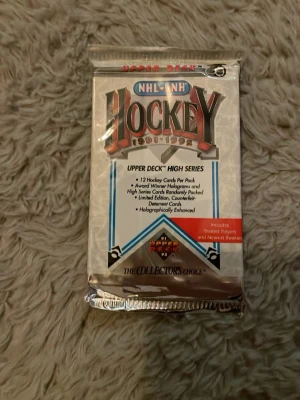 Upper Deck NHL Hockeykort 1991-92 - Oöppnat paket med 12 hockeykort från Upper Deck NHL 1991-1992 High Series. Förpackningen är silverfärgad med blå och röda detaljer och innehåller samlarkort med hologram och specialkort. Perfekt för dig som gillar sport och samlarprylar.