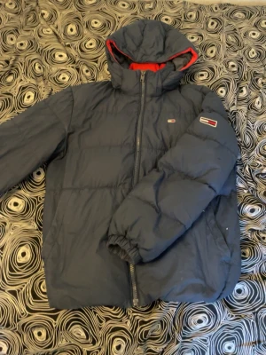 Mörkblå pufferjacka från Tommy Hilfiger - Snygg mörkblå pufferjacka från Tommy Hilfiger med huva och röda detaljer på insidan. Jackan har dragkedja framtill, två sidofickor och elastiska muddar vid ärmsluten. Klassisk logga på bröstet och ärmen. Perfekt för kalla vinterdagar. Köpt för 2000kr använder inte den eftersom den har blivit för liten för mig. oxå att den har hål som visas på bild 7 ☺️