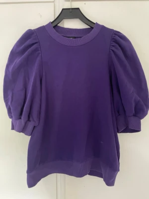 Lila topp med puffärmar från Lindex - Trendig lila topp från Lindex med voluminösa puffärmar och ribbade muddar vid ärmslut och halsringning. T-shirten har en loose passform och är tillverkad i mjuk bomull, perfekt för en cool och avslappnad stil.