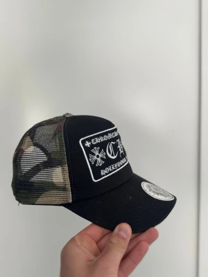 Svart och camo truckerkeps Chrome Hearts - Snygg truckerkeps från Chrome Hearts med svart front och broderad vit logga. Baksidan är i camouflagemönstrat meshmaterial för extra ventilation. Böjd skärm och klassisk snapback-stängning. Perfekt för en streetwear-look.