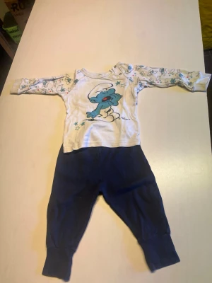 Smurfarna set med långärmad body - Sött set med långärmad body i vitt med Smurfarna-tryck och matchande blå byxor. Bodyn har mönstrade ärmar med små smurfar och tryckknappar på axeln. Byxorna är mjuka och har bred mudd i midjan och benslut. Perfekt för små barn som gillar Smurfarna.