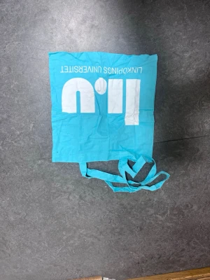 Turkos tygkasse från Linköpings universitet - Turkos shoppingväska i tyg med vitt tryck från Linköpings universitet. Väskan har långa handtag och är perfekt för att bära böcker eller annat. Tryck på ena sidan med universitetets logga och på andra sidan webbadress.
