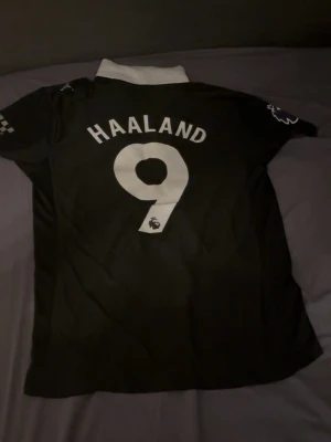 Svart Manchester City Haaland tröja - Säljer en svart Manchester City fotbollströja från Puma med Haaland och nummer 9 på ryggen. Tröjan har vit krage, Premier League-logga på ärmen och Etihad Airways som sponsor framtill. Perfekt för dig som älskar fotboll och vill stötta City. Aldrig använd tröja. Skick 10/10. 