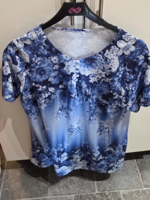 Blå blommig blus med kort ärm XL - Snygg blus i blått med blommigt mönster och korta ärmar. Blusen har rund halsringning och små knappar framtill. Perfekt för dig som gillar färg och mönster. Tillverkad i mjukt och lätt material som känns skönt mot huden.