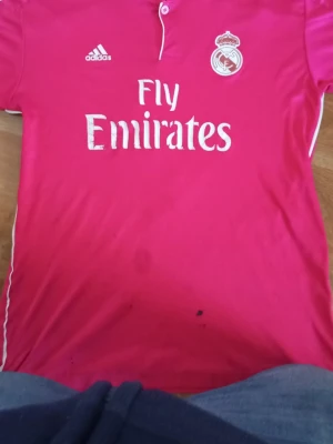 Rosa Real Madrid tröja Ronaldo 7 - Säljer en rosa Real Madrid matchtröja från Adidas med tryck 'Fly Emirates' på framsidan och 'Ronaldo 7' på ryggen. Tröjan har vita detaljer och klubbens emblem på bröstet. Perfekt för fotbollsfans som vill sticka ut på planen eller läktaren. Har köpt flera andra och den e lite stor men den e super bra och tyger e perfekt 