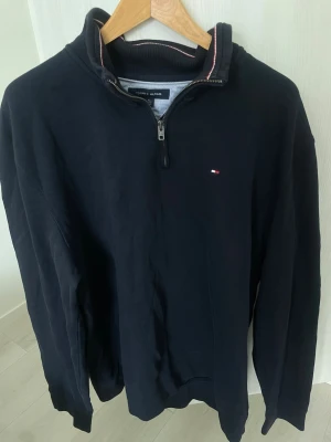 Half zip tröja Tommy Hilfiger - Svart långärmad topp från Tommy Hilfiger med half zip och liten broderad logga på bröstet. Ståkrage med diskret röd och vit rand på insidan. Tillverkad i mjuk bomull för en clean och stilren look.
