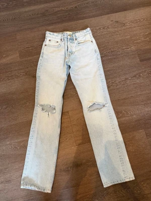 Ljusblå raka jeans med slitningar - Säljer ett par ljusblå jeans med raka ben och slitna detaljer vid knäna. Jeansen har klassisk femficksdesign och hög midja. Perfekta för en avslappnad och trendig look. Materialet är denim i bomull.