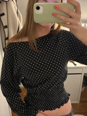 Svart prickig croppad topp - Svart långärmad topp med vita prickar och croppad passform. Toppen har smockad midja och vågig kant nertill, vilket ger en snygg siluett. Perfekt för dig som gillar trendiga och bekväma plagg.
