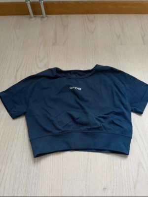 Mörkblå cropped t-shirt från DFYNE - Snygg mörkblå cropped t-shirt från DFYNE med korta ärmar och rund halsringning. T-shirten har en tight passform och logga både fram och bak. Perfekt för träning eller gymmet. Materialet är stretchigt och andas bra.