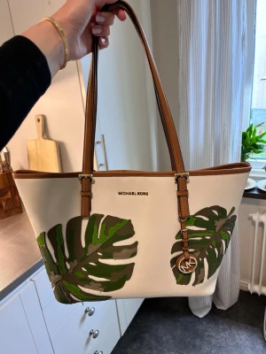 Michael Kors handväska med bladmotiv - Snygg handväska från Michael Kors i vit canvas med bruna läderhandtag och gröna bladmotiv på framsidan. Väskan har en klassisk tote-form och MK-hänge i metall. Perfekt för dig som gillar trendiga detaljer och naturinspirerat mönster. Köpt i Köln tror jag för 4500kr