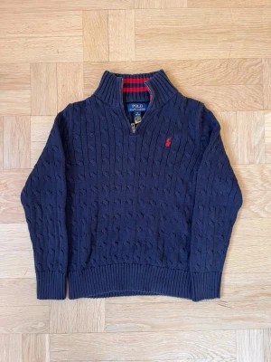 Mörkblå kabelstickad tröja Polo Ralph Lauren - Kabelstickad mörkblå tröja från Polo Ralph Lauren med halv dragkedja och röd broderad logga på bröstet. Tröjan har ribbade muddar och krage med röd-rutigt foder. Perfekt för dig som gillar klassisk och stilren look.