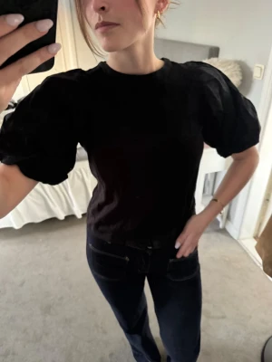 Svart topp med puffärmar - Svart topp med trendiga puffärmar och rund halsringning. Modellen är croppad och har en loose passform som ger en cool siluett. Perfekt att styla med jeans eller kjol för en snygg look.