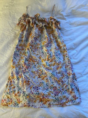Blommig sommarklänning med volang - Söt klänning med tunna axelband och volang upptill. Mönstret är blommigt i lila, orange, grönt och gult på vit botten. Klänningen är luftig och har en rak passform, perfekt för varma dagar. Materialet känns lätt och mjukt, troligen bomull eller viskos.