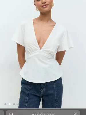 Vit omlottopp från Gina Tricot - Vit omlottopp från Gina Tricot med djup v-ringning och korta ärmar. Toppen har en figurnära passform med knyt i midjan och är tillverkad i mjuk bomull. Perfekt att styla med jeans eller kjol.