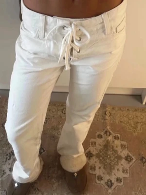 Vita jeans med snörning i midjan - Säljer ett par vita/krämfärgade jeans med unik snörning framtill i midjan. Jeansen har raka ben och klassiska fickor fram och bak. Snygg och trendig modell som sticker ut med sitt coola snörningsdetalj. Perfekta för dig som vill ha något annorlunda i garderoben.