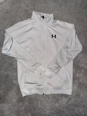 Vit zip-hoodie från Under Armour - Snygg vit hoodie från Under Armour med dragkedja hela vägen och hög krage. Tröjan har två fickor framtill och svart logga på bröstet. Tillverkad i mjukt och sportigt material, perfekt för en aktiv livsstil.