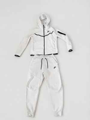 Nike Tech Fleece vit loungewear-set - Nike Tech Fleece loungewear-set i vitt med svarta detaljer. Setet består av en hoodie med dragkedja, fickor och svarta kanter samt matchande byxor med dragsko och muddar. Båda plaggen har Nike-logga och är tillverkade i mjukt fleecematerial.