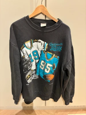 Svart Jacksonville Jaguars sweatshirt - Cool svart sweatshirt med stort tryck av Jacksonville Jaguars på framsidan. Motivet visar två matchtröjor, hjälm och amerikansk fotboll i blått, vitt och gult. Tröjan har rund hals och ribbade muddar. Perfekt för dig som gillar NFL och sportig stil.