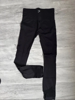 Svarta vital seamless leggings Gymshark - Svarta seamless leggings från Gymshark med hög midja och diskreta meshdetaljer längs benen. Stretchigt material som sitter tight och bekvämt, perfekt för träning eller chill. Gymshark-logga framtill vid midjan.