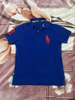 Blå pikétröja från Ralph Lauren - Snygg blå pikétröja från Ralph Lauren med röd broderad logga på bröstet och siffra på ärmen. Klassisk krage med knappar och gröna detaljer på insidan. Perfekt för en stilren och sportig look.