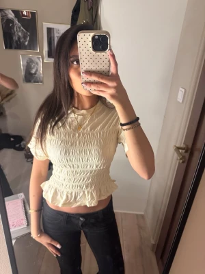 Ljusgul t Shirt  - Säljer en ljusbeige croppad blus med korta ärmar och smockad struktur. Toppen har volang nertill och är perfekt för dig som gillar en feminin och trendig stil. Passformen är normal och materialet känns som bomull. Köpt och enbart testat men passade inte mig😊