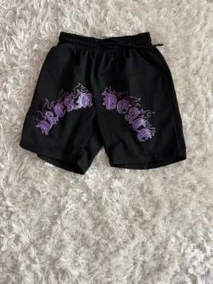 Svarta shorts med lila tryck - Coola svarta shorts med elastisk midja och stort lila tryck framtill där det står 'DREAM PEACE' i gotisk stil med flames. Perfekta för en avslappnad streetwear-look.