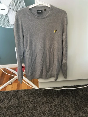 Grå tröja från Lyle & Scott - Snygg grå långärmad tröja från Lyle & Scott med rund halsringning och klassisk gul logotyp på bröstet. Tröjan är i mjukt och tunt stickat material, perfekt för lager-på-lager och enkel att matcha med jeans eller chinos. Lite utdragen i halsen men funkar bra ändå! 