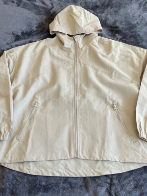 Alo Yoga Vantage Nylon Ripstop Track Jacket Oversized  - Stilren beige vindjacka med dragkedja framtill och två snedställda fickor med dragkedja. Jackan har en rymlig huva och är tillverkad i ett lätt material som passar perfekt för blåsiga dagar. Enkel och clean design som funkar till många outfits.