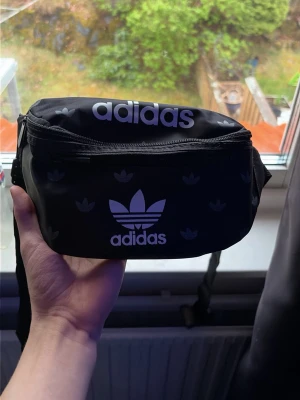 Svart magväska från Adidas - Snygg svart magväska från Adidas med vita loggor och justerbart band. Väskan har två fack med dragkedja och är tillverkad i slitstarkt syntetmaterial. Perfekt för att bära det viktigaste när du är på språng.