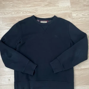 Svart sweatshirt från Burberry - Klassisk svart sweatshirt från Burberry med rund halsringning och diskret broderad logga på bröstet. Tillverkad i mjukt material och har ribbade muddar vid ärmslut och nederkant. Insidan har det ikoniska Burberry-mönstret i rött och grått.