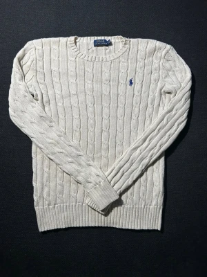 Vit kabelstickad tröja Polo Ralph Lauren - Kabelstickad tröja i offwhite från Polo Ralph Lauren med rund halsringning och blå broderad logga på bröstet. Tröjan har ribbade muddar vid ärmslut och nederkant och är tillverkad i mjukt material. Perfekt för lager på lager och klassisk stil.