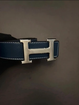 Blått skärp med H-spänne - Snyggt blått skärp med ikoniskt H-format spänne i silverfärgad metall. Skärpet är gjort i skinn med vita kontrastsömmar längs kanterna. Perfekt accessoar för att lyfta din outfit.