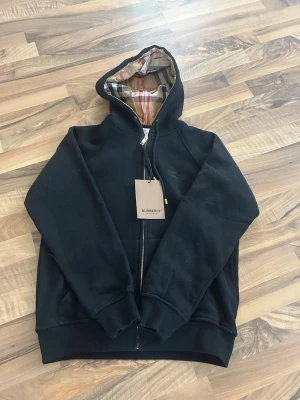  Burberry zip up  - Snygg svart hoodie från Burberry med dragkedja och klassiskt rutigt foder i huvan. Broderad Burberry-logga på bröstet och ribbade muddar vid ärmar och nederkant. Perfekt för dig som vill ha en stilren och exklusiv look.