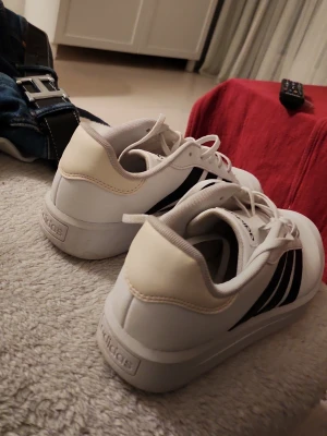 Vita Adidas sneakers med svarta ränder - Snygga vita Adidas sneakers med klassiska svarta ränder på sidorna och vit sula. Skorna har lågt skaft och är tillverkade i syntetmaterial med textilfoder. Perfekta för en clean och sportig look.