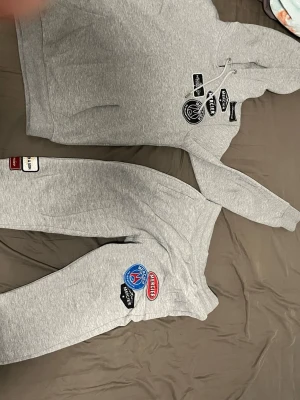 Grått PSG loungewear-set med patches - Säljer ett grått loungewear-set med hoodie och byxor. Setet har flera coola patches och tryck, bland annat PSG-logga och andra märken på både tröjan och byxorna. Hoodien har dragsko och stor ficka framtill. Perfekt för chill dagar hemma.