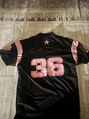 Svart Arizona tröja med rosa detaljer - Snygg svart sporttröja från Colosseum med rosa detaljer och tryck. Stort nummer 36 på både fram- och baksida, samt texten ARIZONA framtill. Rosa stjärna på ryggen och tryck på ärmarna. Perfekt för dig som gillar amerikansk sportstil.