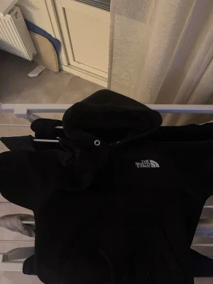 Svart hoodie från The North Face - Säljer en svart hoodie från The North Face med klassisk logga på bröstet. Hoodien har en stor huva med dragsko och en magficka framtill. Perfekt för dig som gillar streetwear och vill ha något bekvämt och stilrent.