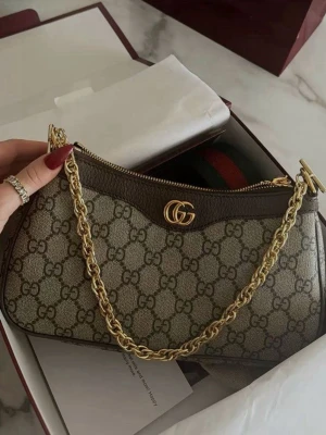 Gucci GG Supreme handväska med kedja - Säljer en snygg handväska från Gucci i GG Supreme canvas med bruna läderdetaljer och guldfärgad kedja. Väskan har det klassiska GG-mönstret och en elegant form med dragkedja upptill. Perfekt accessoar för dig som gillar lyxig och trendig stil.