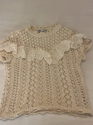 Beige stickad topp med volang Zara - Söt beige stickad topp från Zara med korta ärmar och dekorativ volang i broderad spets över bröstet. Tröjan har ett fint hålmönster och ribbad halsringning. Perfekt för en trendig och romantisk look.