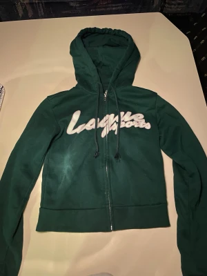 Grön hoodie från Divided H&M XS - Mörkgrön hoodie med dragkedja från Divided  i storlek XS. Trycket 'League Sports' i vitt framtill ger en sportig vibe. Hoodien har huva med dragsko och är tillverkad i mjukt sweatshirtmaterial. Perfekt för en avslappnad look.