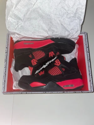 Air Jordan 4 Retro Svart/Röd - Säljer ett par Air Jordan 4 Retro sneakers i svart och rött. Skorna har meshpaneler, mockadetaljer och klassiska plastdetaljer vid snörningen. Sulan är tjock och har synlig Air-enhet. Perfekta för dig som vill sticka ut med en ikonisk streetwear-look.