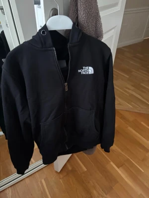 Svart kofta från The North Face - Säljer en svart hoodie från The North Face med dragkedja och huva. Klassisk logga på bröstet och två stora fickor framtill. Perfekt för en chill och stilren look. Mjuk bomullskänsla och ribbade muddar vid ärmslut och nederkant. Storlek S.