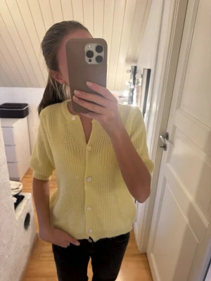 Gul stickad kortärmad kofta - Söt ljusgul stickad kofta med korta ärmar och rund halsringning. Koftan har ribbstickad struktur och knäpps med vita knappar framtill. 