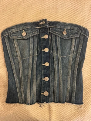 Jeanskorsett med knappar framtill - Säljer en snygg jeanskorsett i blå denim med dekorativa sömmar och fem silverfärgade knappar framtill. Korsetten har två bröstfickor med lock och knapp samt smockad rygg för bättre passform. Rå kant nedtill för en trendig look.