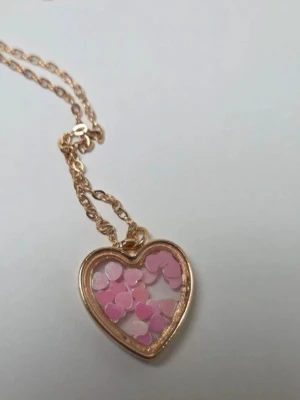 Halsband med hjärthänge och rosa detaljer - Sött halsband med ett hjärtformat hänge i guldfärgad metall. Hänget har en transparent mitt fylld med små rosa hjärtformade dekorationer som ger en lekfull och trendig look. Perfekt accessoar för dig som gillar gulliga och unika smycken.