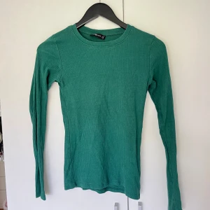 Grön ribbad långärmad tröja - En grön långärmad tröja från i ribbad bomull. Tröjan har rund halsringning och en slim passform som sitter snyggt på kroppen. Perfekt att styla med jeans eller kjol för en chill look.