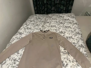 Beige henley från Patagonia - Beige henley med lång ärm från Patagonia. Tröjan har tre knappar vid halsen och en liten logotyp på bröstet. Tillverkad i mjuk polyester och har en clean, enkel design som funkar till det mesta.