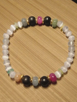 Färgglatt pärlarmband med gulddetaljer - Snyggt armband med pärlor i olika färger som vitt, svart, rosa, grön och blå. Armbandet har små guldfärgade detaljer mellan vissa pärlor och är elastiskt för enkel användning. Perfekt accessoar för att addera färg till din stil. Pärlorna är stenpärlor.  Armbandet är elastiskt.