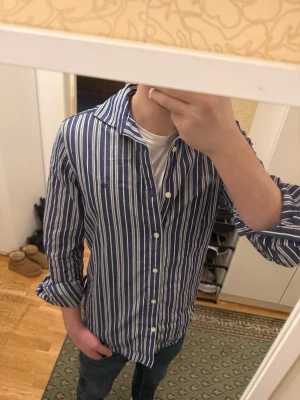 Blåvit randig skjorta från Morris - Snygg blå skjorta från Morris med vita vertikala ränder och klassisk krage. Tillverkad i bomullstyg från Tessitura Monti. Skjortan har långa ärmar och knäppning framtill. Perfekt för en stilren och avslappnad look.